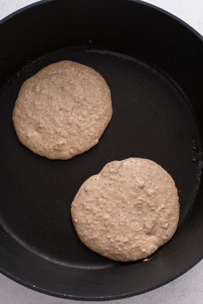 Two oatmeal pancakes being cooked in a cast iron skillet.