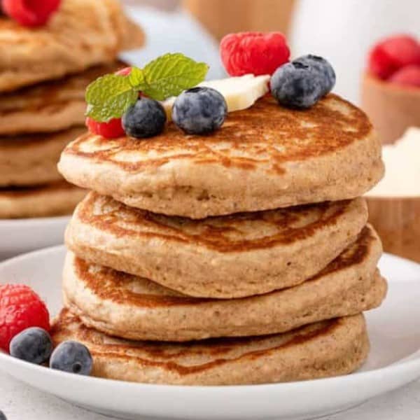 White plate with a stack of four oatmeal pancakes. A second stack of pancakes and container of syrup are in the background.