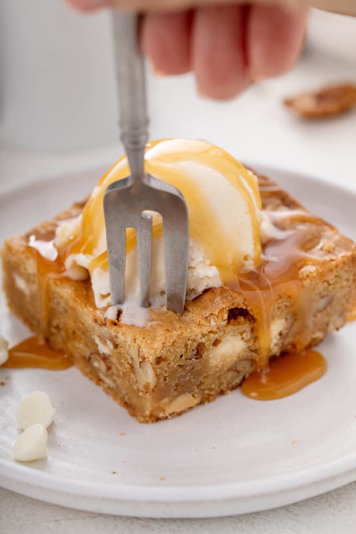 Fork about to take a bite from a plated white chocolate pecan blondie that's topped with ice cream and caramel sauce.