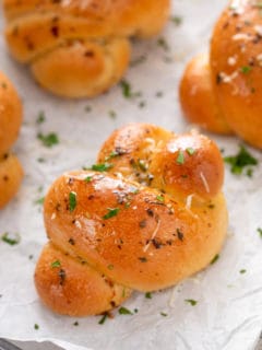 Close up of garlic knots on a piece of parchment paper, topped with parmesan cheese and fresh parsley.