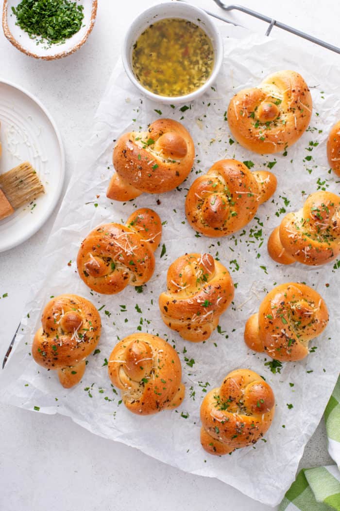 Overhead view of baked garlic knots on a piece of parchment paper, garnished with parsley and parmesan cheese.