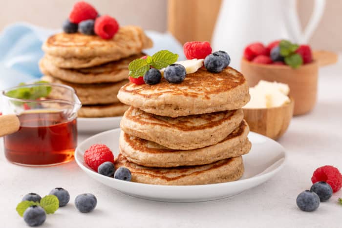 White plate with a stack of four oatmeal pancakes. A second stack of pancakes and container of syrup are in the background.
