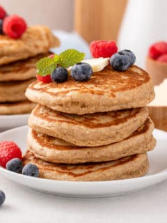 Four oatmeal pancakes stacked on a white plate and topped with butter and berries.