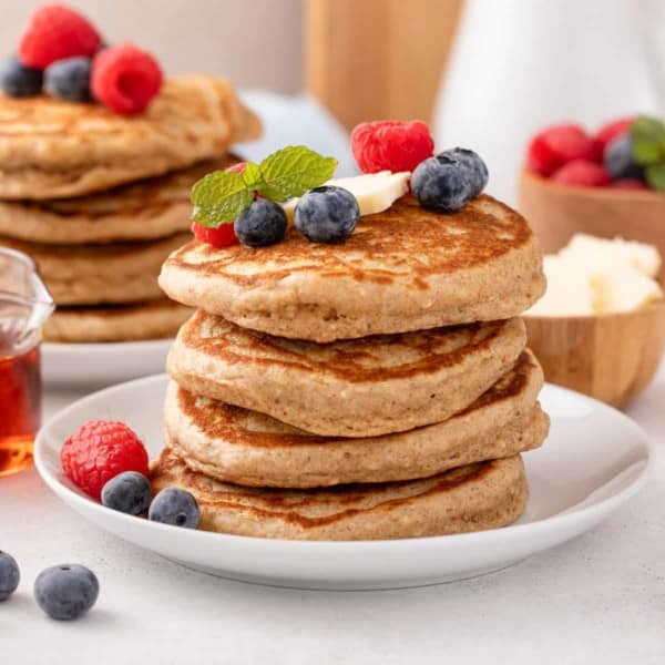Four oatmeal pancakes stacked on a white plate and topped with butter and berries.