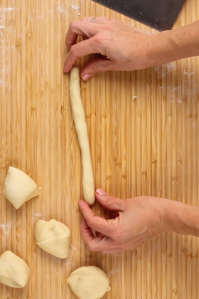 Hands rolling a piece of dough into a snake to make into a garlic knot.