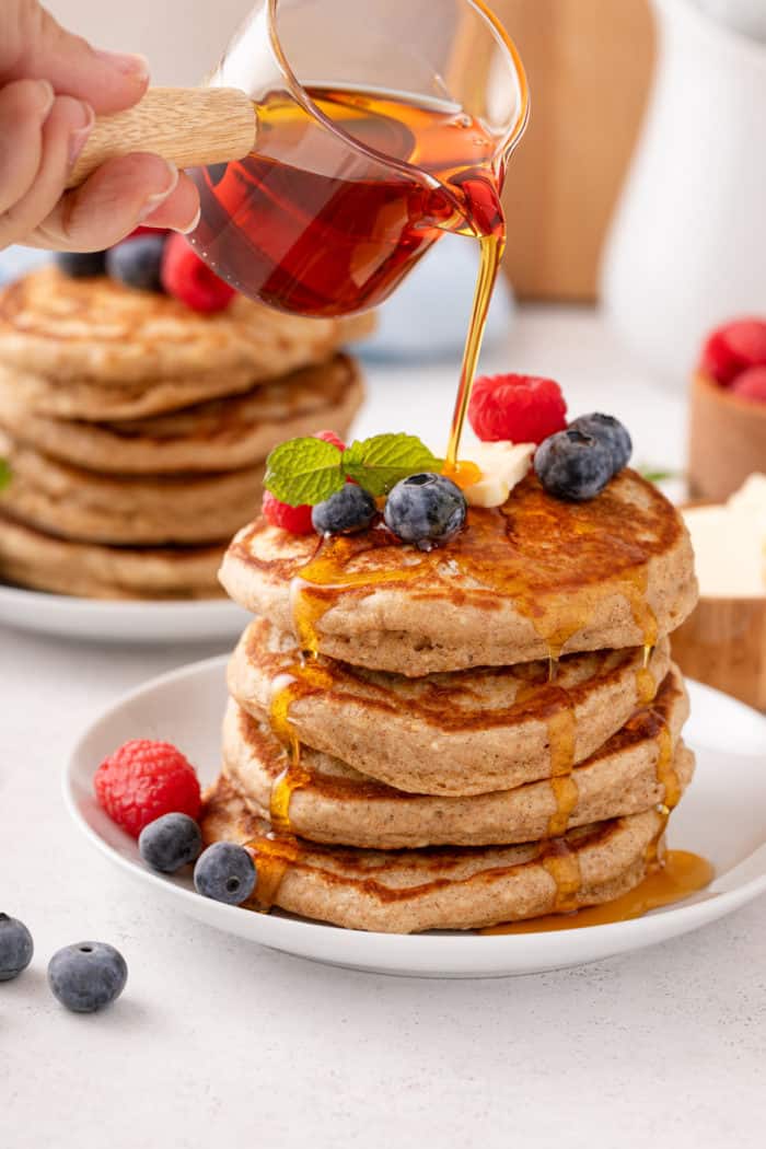 Syrup being poured over a stack of oatmeal pancakes topped with butter and fresh berries.