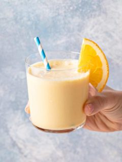 Hand holding up a glass of orange julius, garnished with a blue and white striped straw and an orange slice.