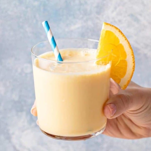 Hand holding up a glass of orange julius, garnished with a blue and white striped straw and an orange slice.