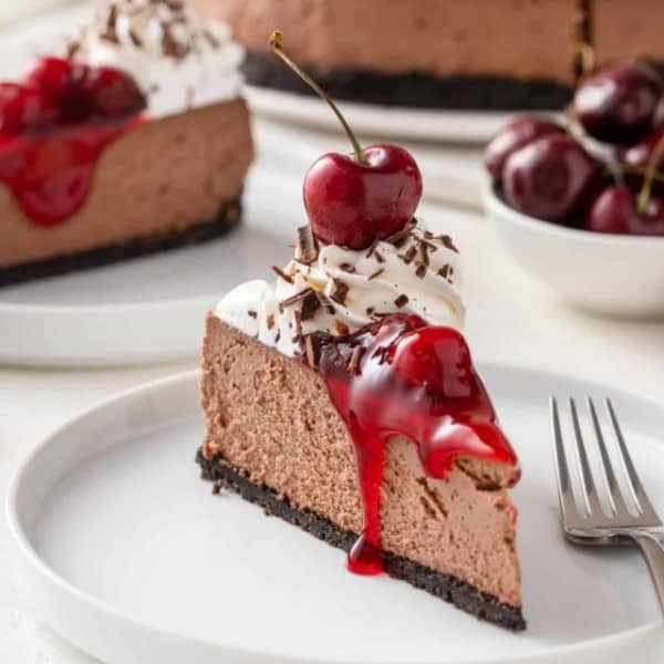 Slice of garnished black forest cheesecake next to a fork on a white plate.