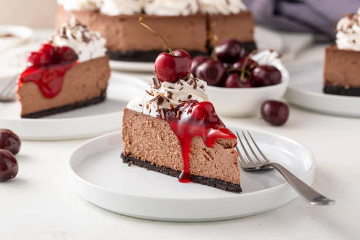 Side view of a slice of black forest cheesecake on a white plate, with additional plates and the whole cheesecake in the background.