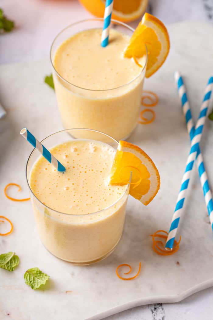 Two glasses of homemade orange julius on a countertop next to blue and white straws.