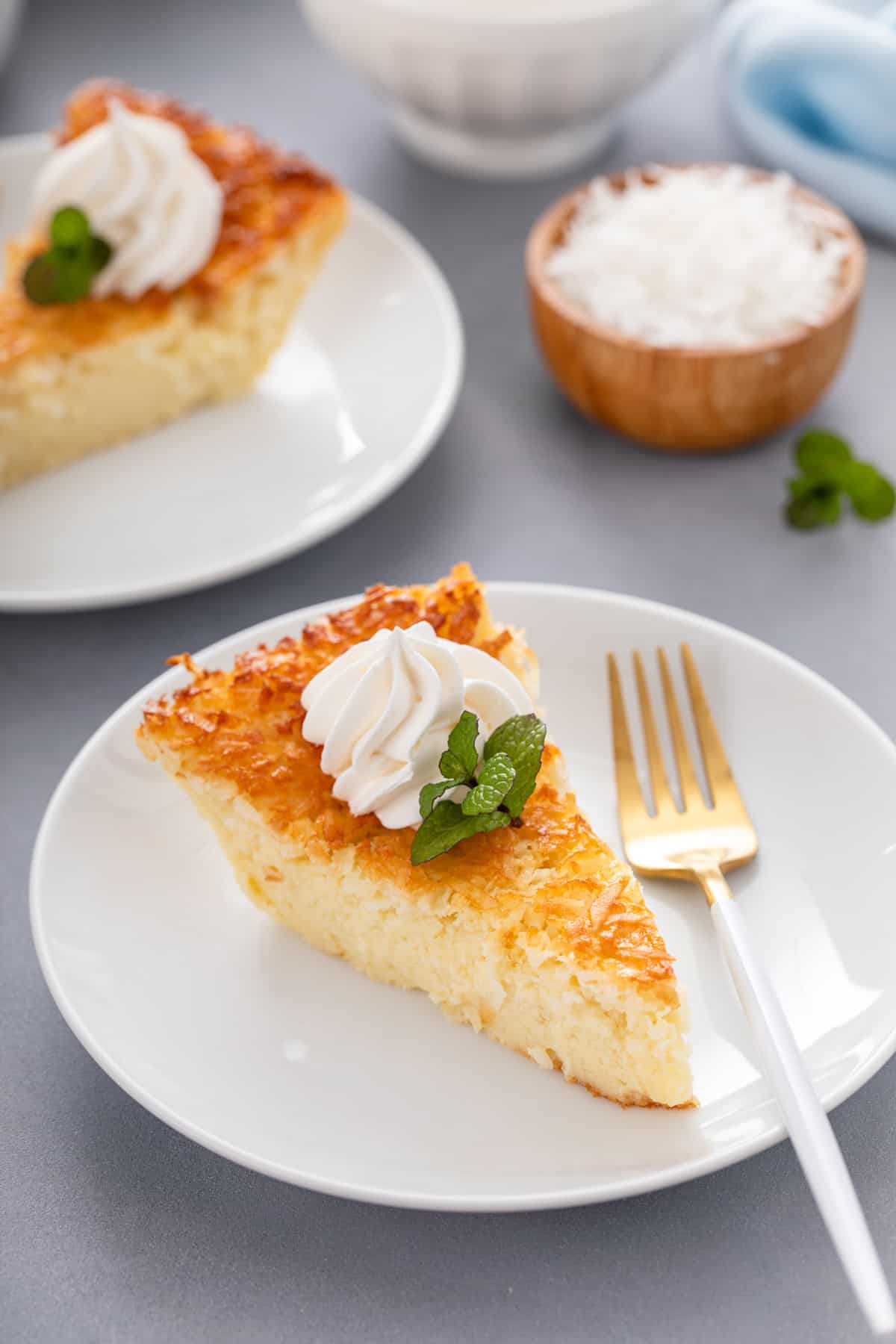 Slice of impossible coconut pie next to a fork on a white plate, garnished with mint and whipped cream.
