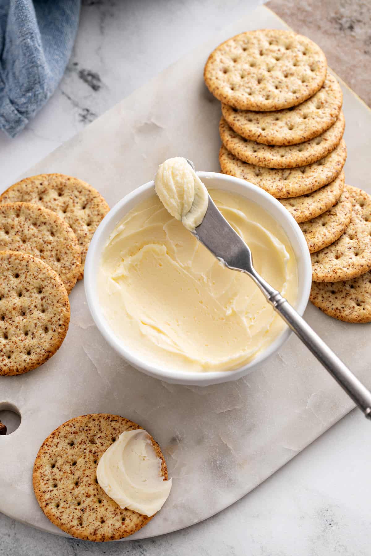 Overhead view of a bowl of butter surrounded by crackers on a marble board. One of the crackers has butter spread onto it.