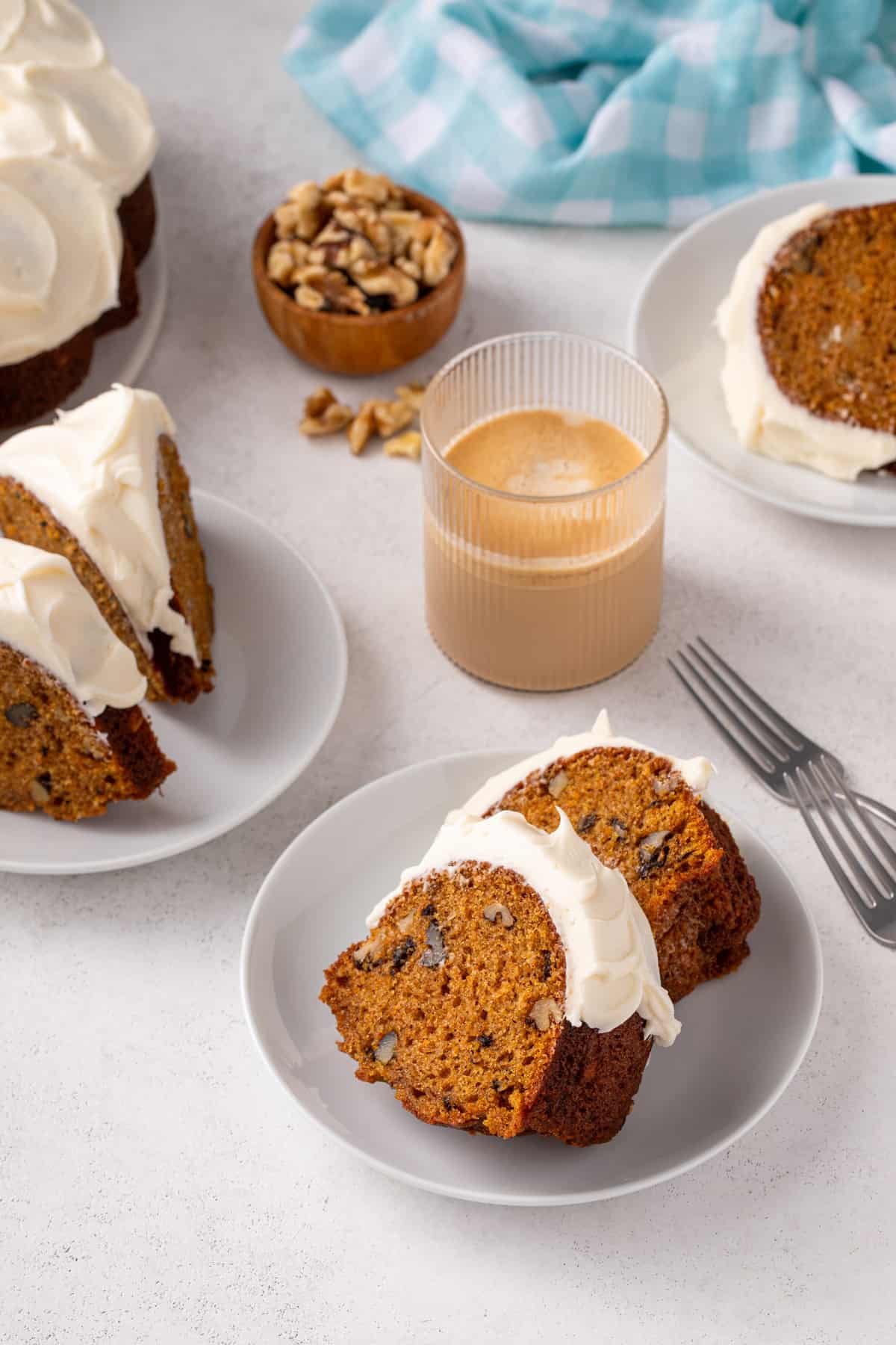 Two plates, each holding two slices of frosted carrot bundt cake, with more cake and iced coffee in the background.