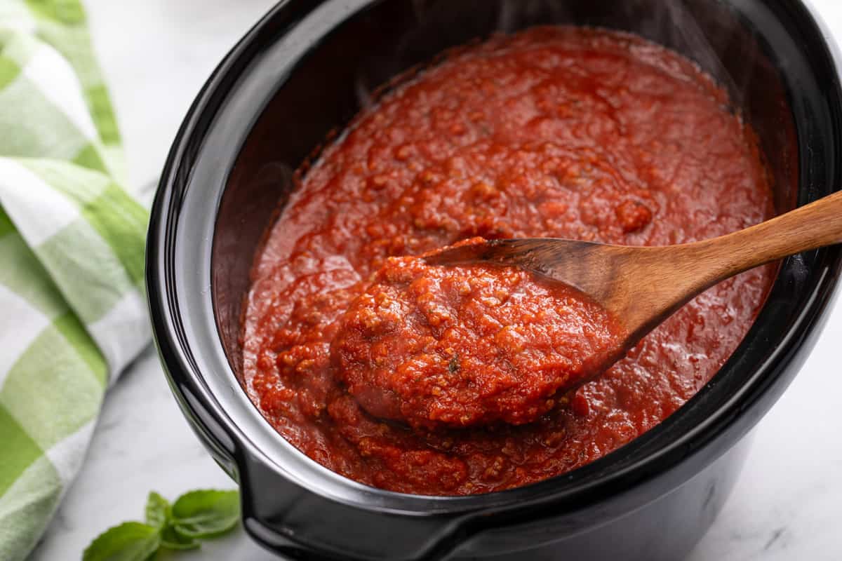 Wooden spoon holding a spoonful of spaghetti sauce over a crockpot filled with the sauce.