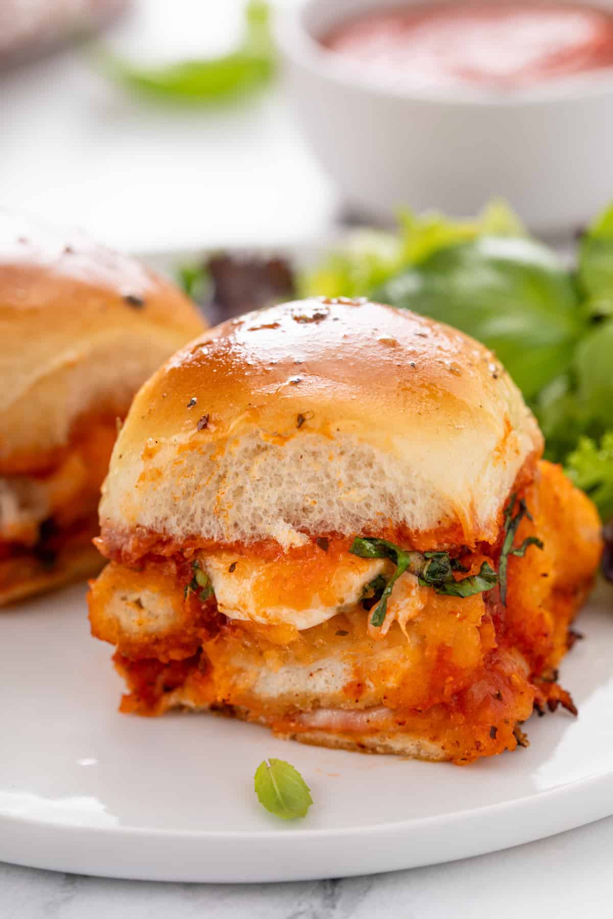 Chicken parmesan slider on a white plate with a green salad in the background.