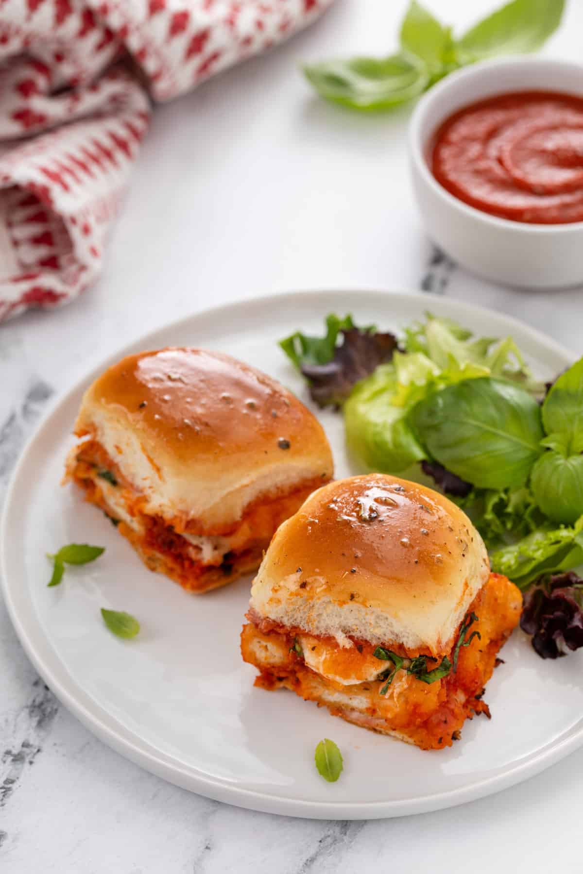 Two chicken parmesan sliders next to a green salad on a white plate.