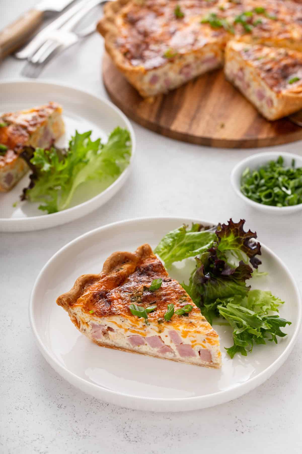 Two plates, each holding a slice of ham and cheese quiche next to fresh greens, with the rest of the quiche in the background.