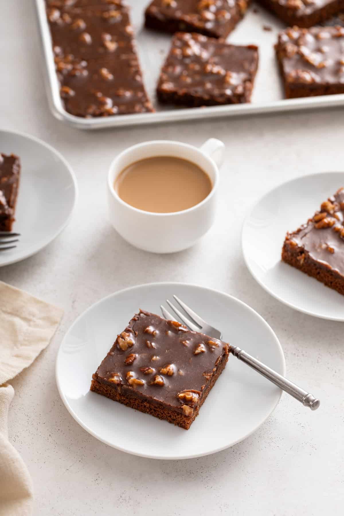 Multiple plates of texas sheet cake next to a cup of coffee. The pan of cake is visible in the background.