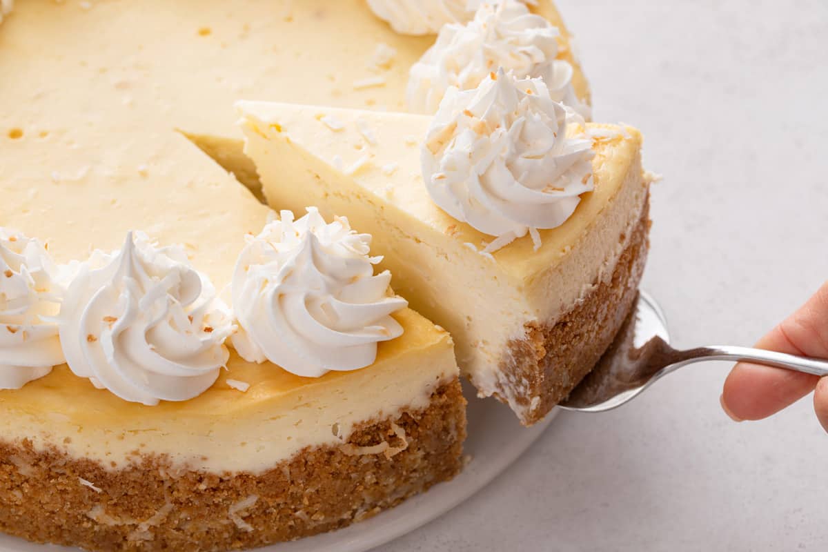 Cake server lifting a slice of coconut cheesecake from a cake plate holding the rest of the cheesecake.