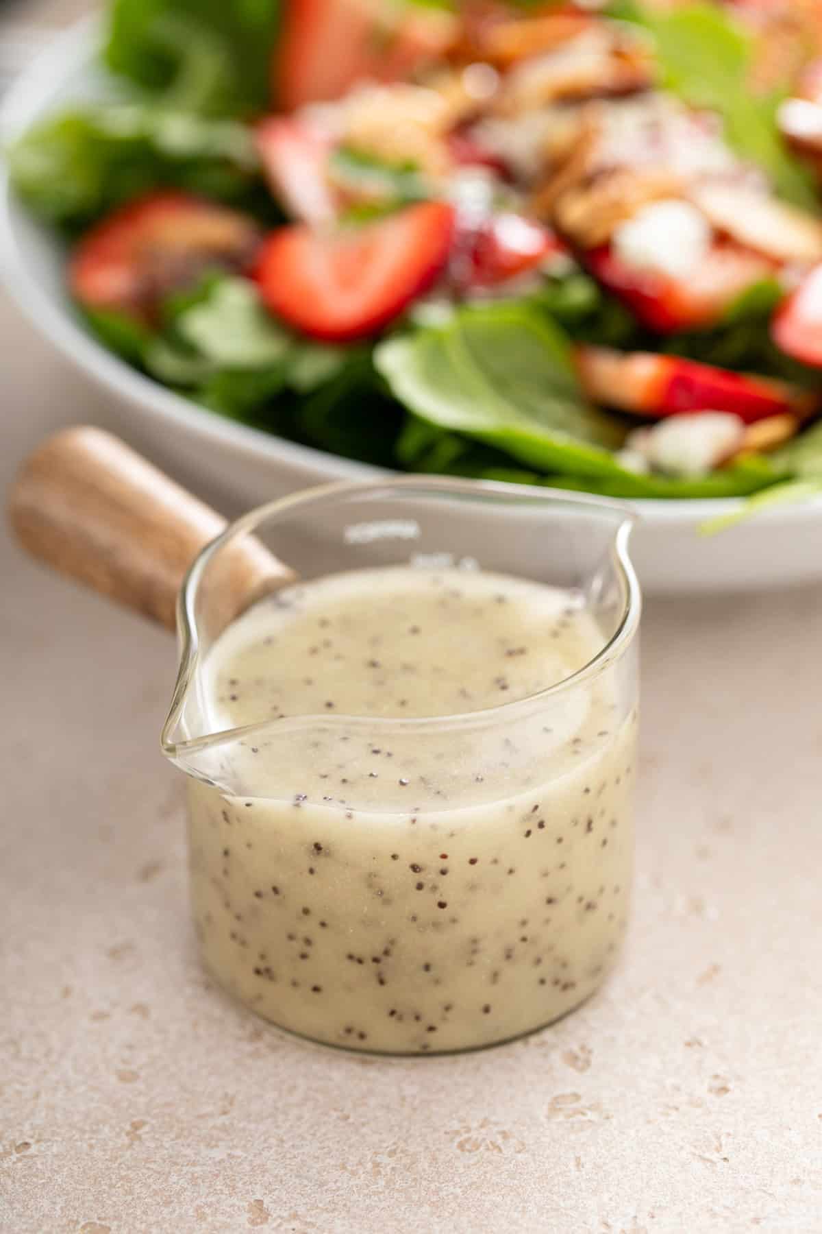 Close up of a small jar of poppy seed dressing with a bowl of strawberry spinach salad in the background.