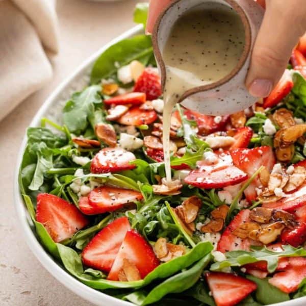 Poppy seed dressing being poured over strawberry salad in a serving bowl.