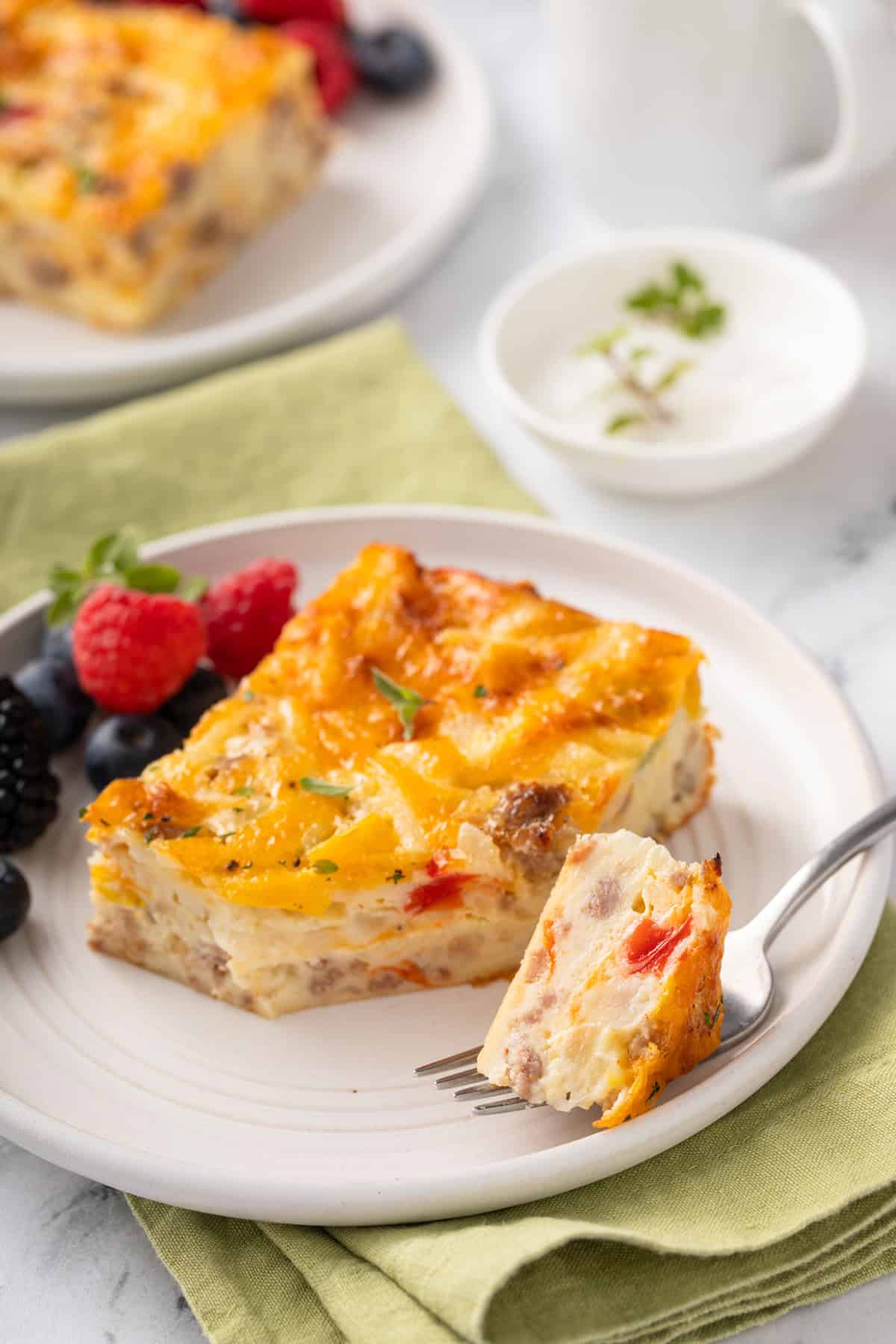 Fork holding a bite of bisquick breakfast casserole next to a slice of casserole on a white plate.