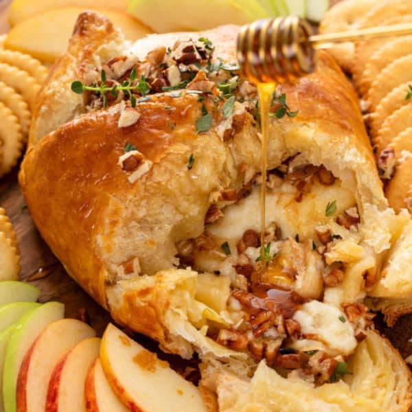 Baked brie in puff pastry that has been cut into on a board with apples and crackers. Honey is being drizzled over the cheese.