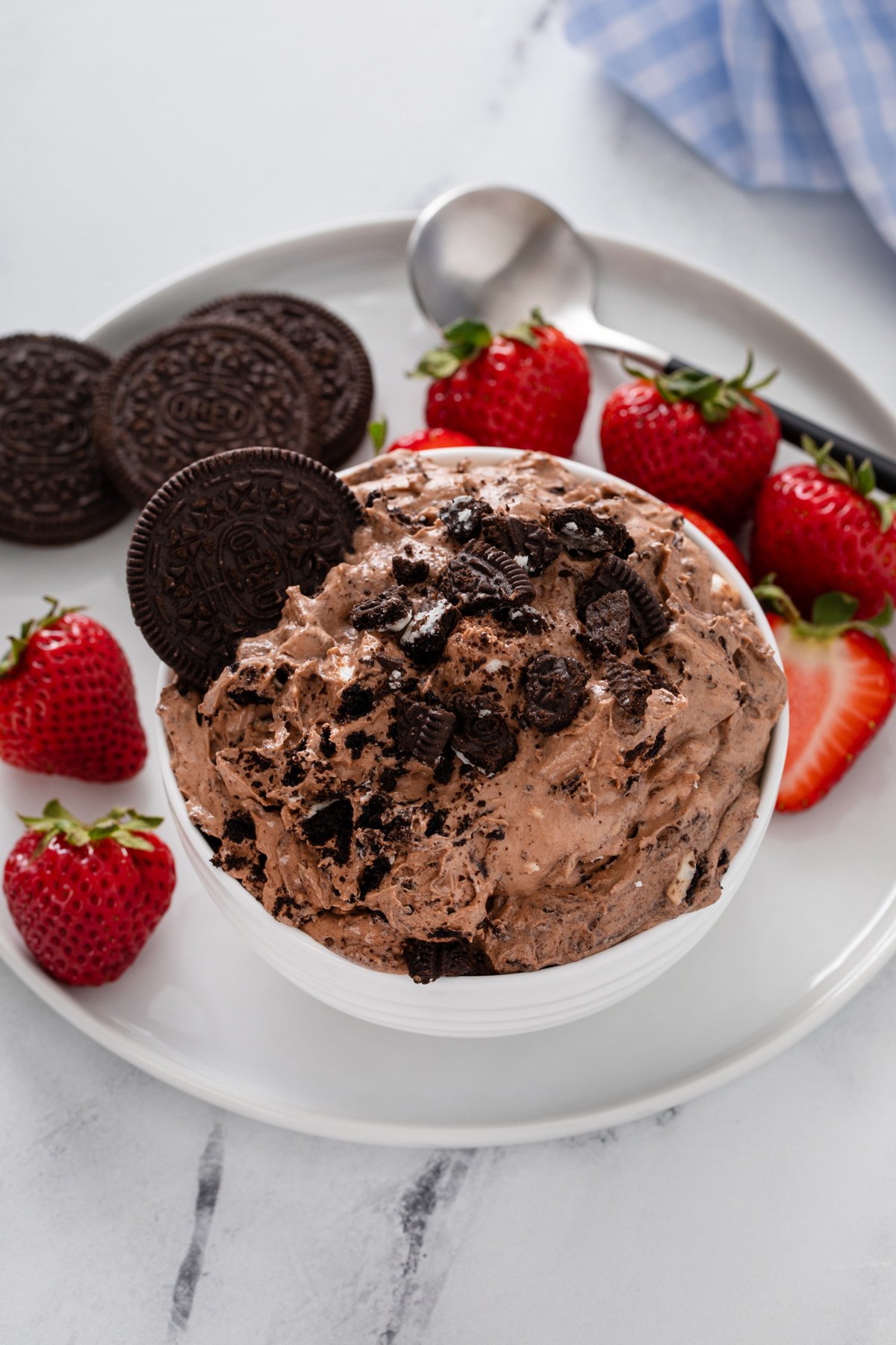 Small white bowl filled with oreo fluff set on a white plate with oreo cookies and fresh strawberries.