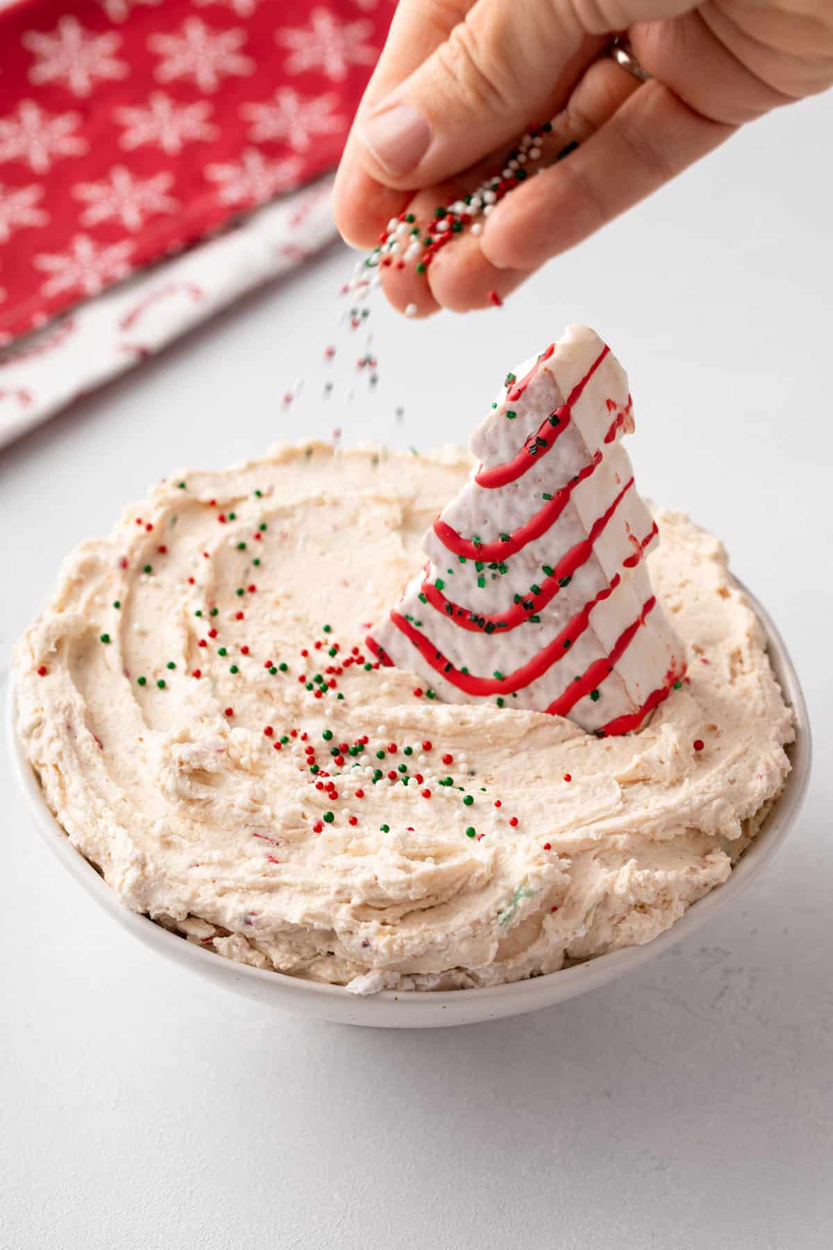 Person sprinkling red and green sprinkles over the top of a bowl of christmas tree cake dip, garnished with a full christmas tree cake.