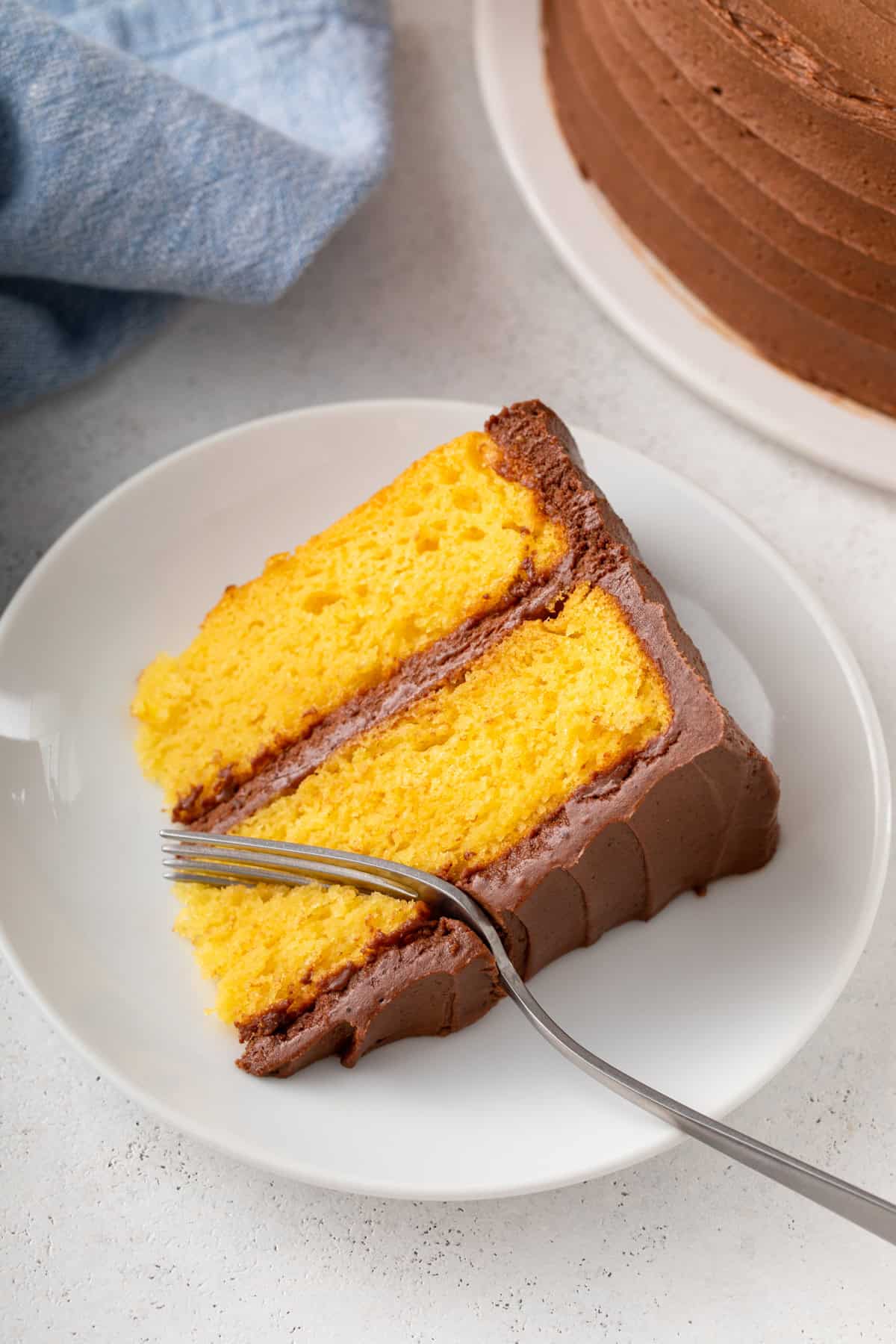 Fork cutting into a slice of yellow layer cake with chocolate frosting on a white plate.