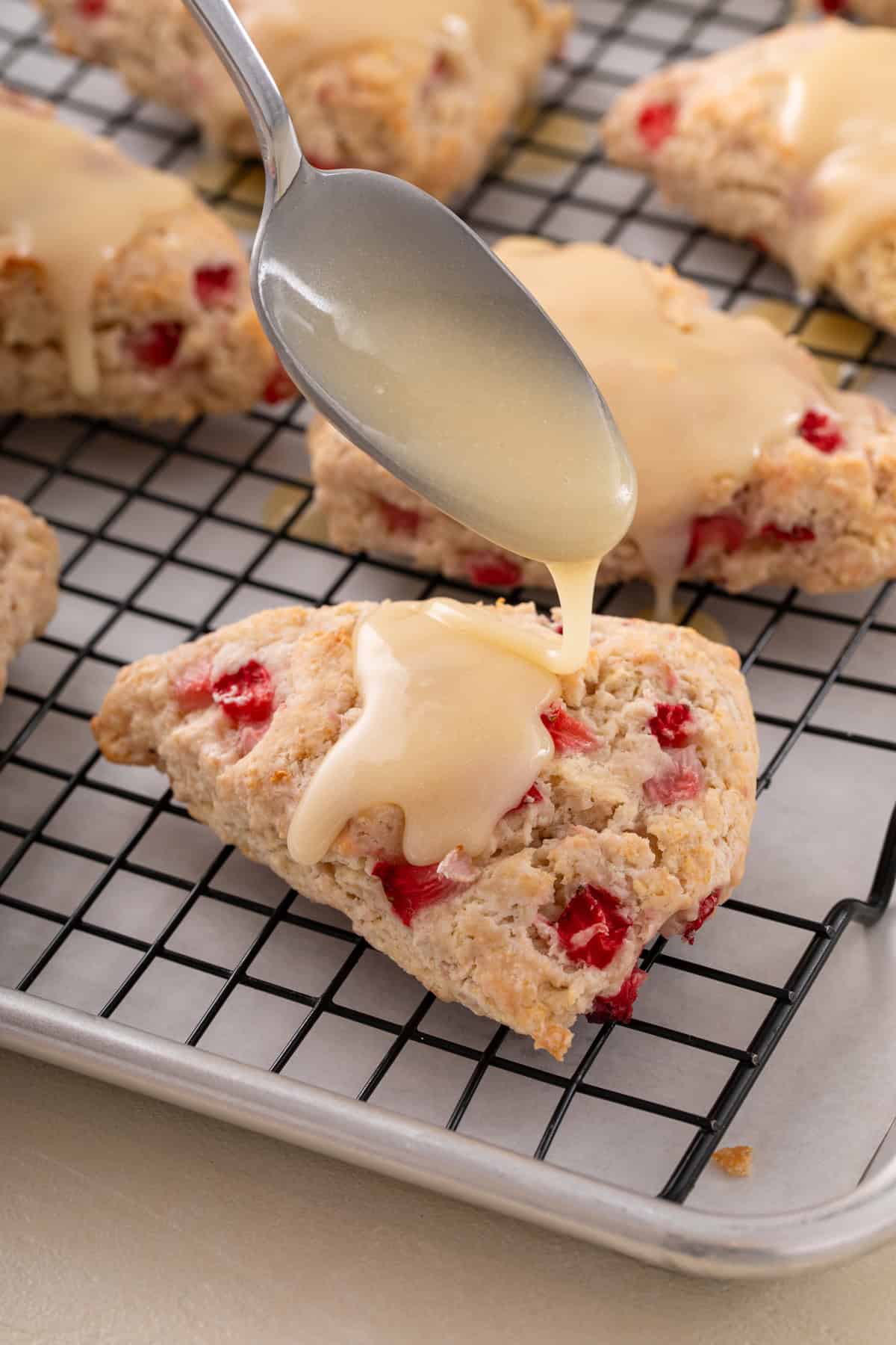 Vanilla glaze being spooned on top of strawberry scones on a wire rack.