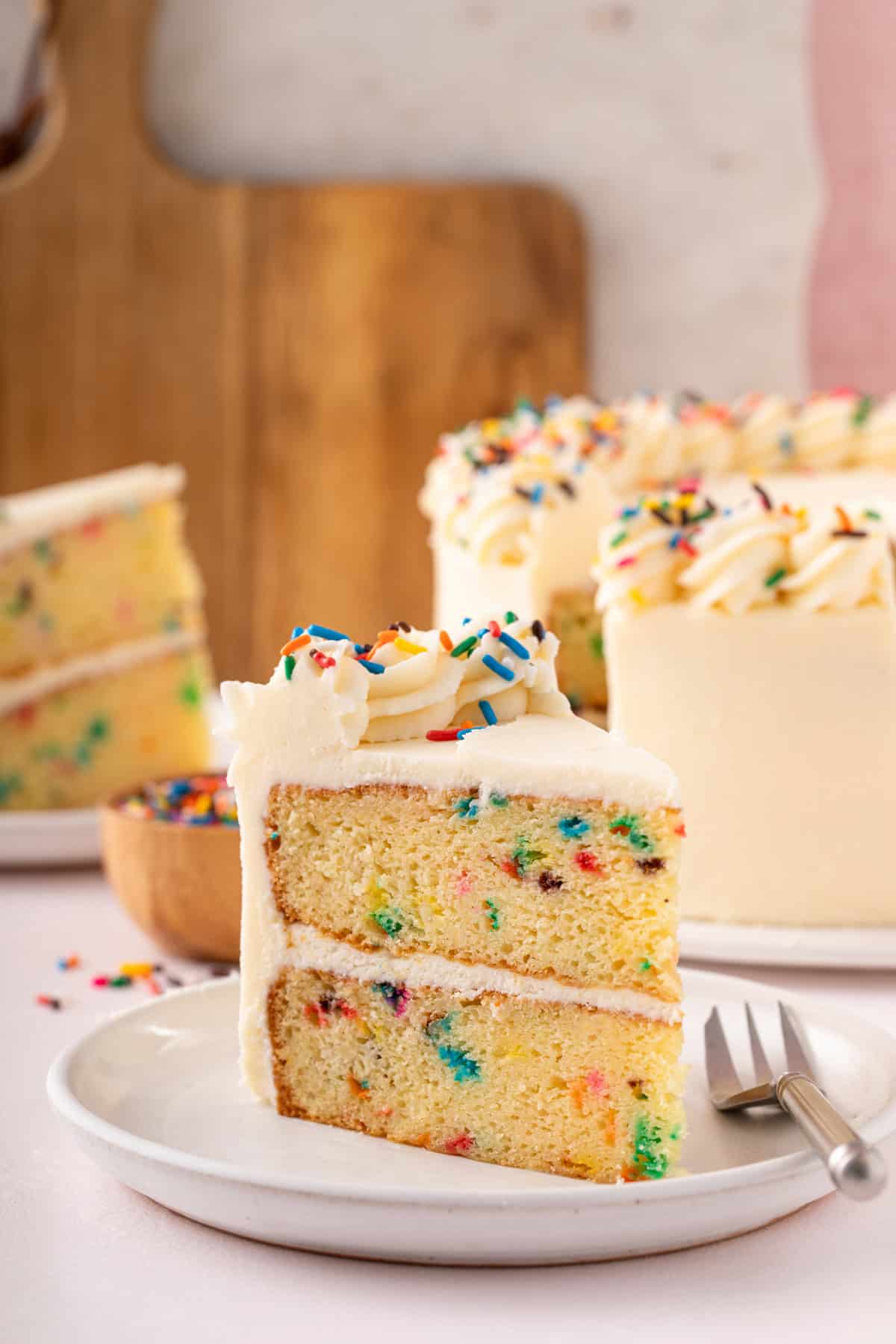 Slice of funfetti layer cake frosted with vanilla frosting and decorated with rainbow sprinkles on a white plate.