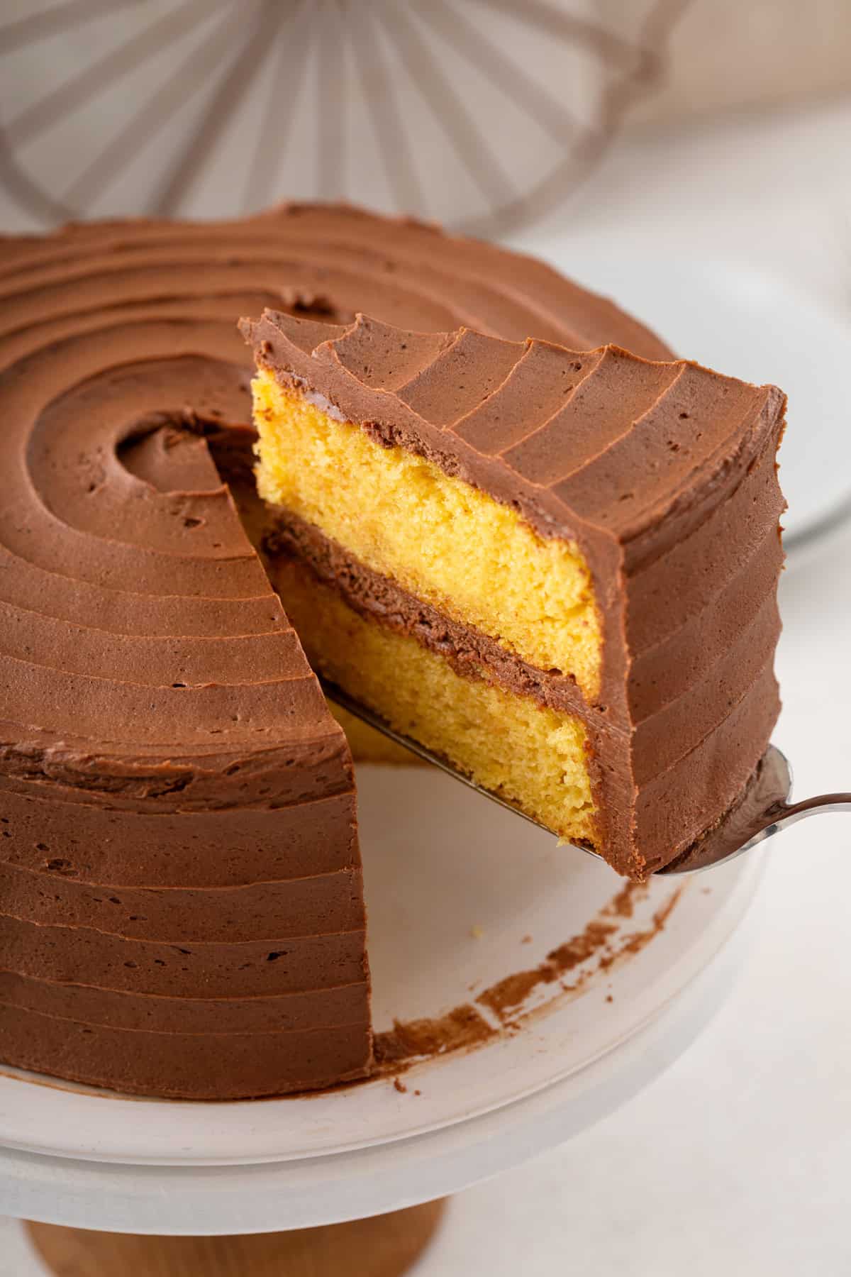 Cake server lifting a slice of easy yellow layer cake topped and filled with chocolate frosting.