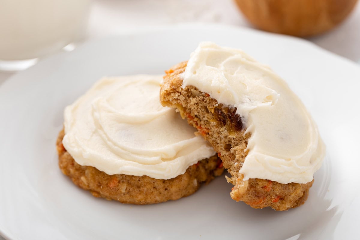 Two frosted carrot cake cookies on a white plate, with a bite taken from one of them.
