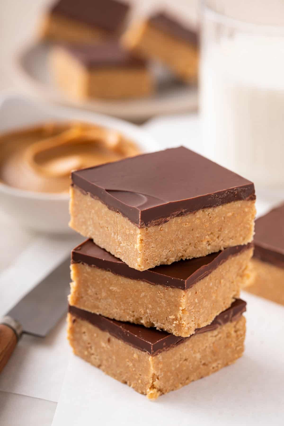 Three lunch lady peanut butter bars stacked on a piece of parchment with a glass of milk and bowl of peanut butter in the background.