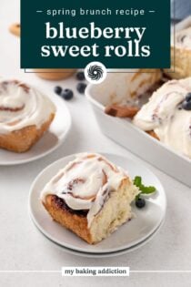 White plate holding a blueberry sweet roll, with a second plate and the pan of rolls in the background. Text overlay includes recipe name.