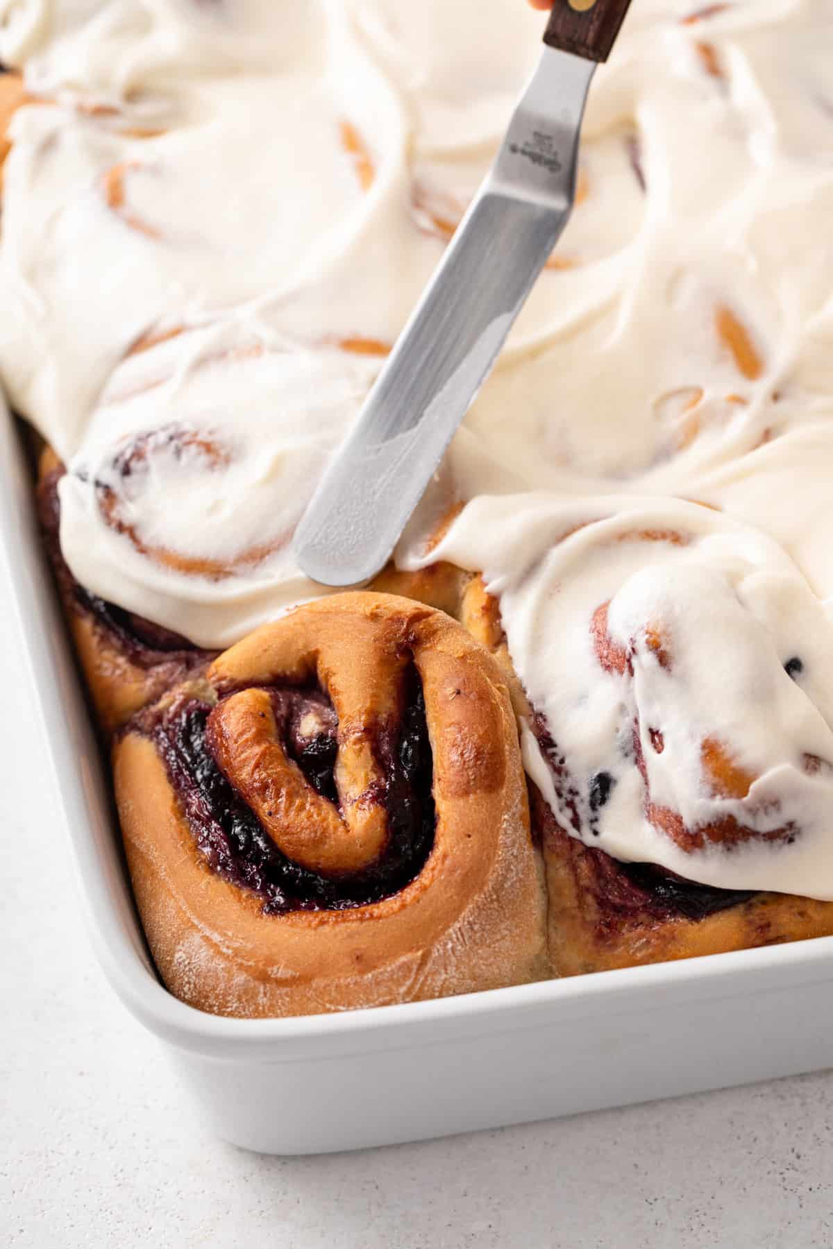 Cream cheese frosting being spread over blueberry sweet rolls in a white baking dish.