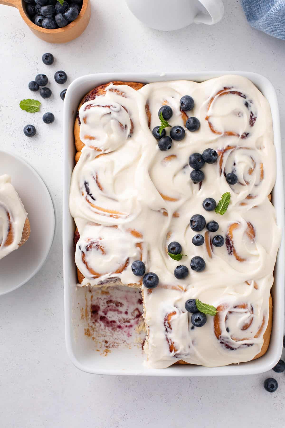 Overhead view of a pan of blueberry sweet rolls with one roll removed from the corner of the pan.