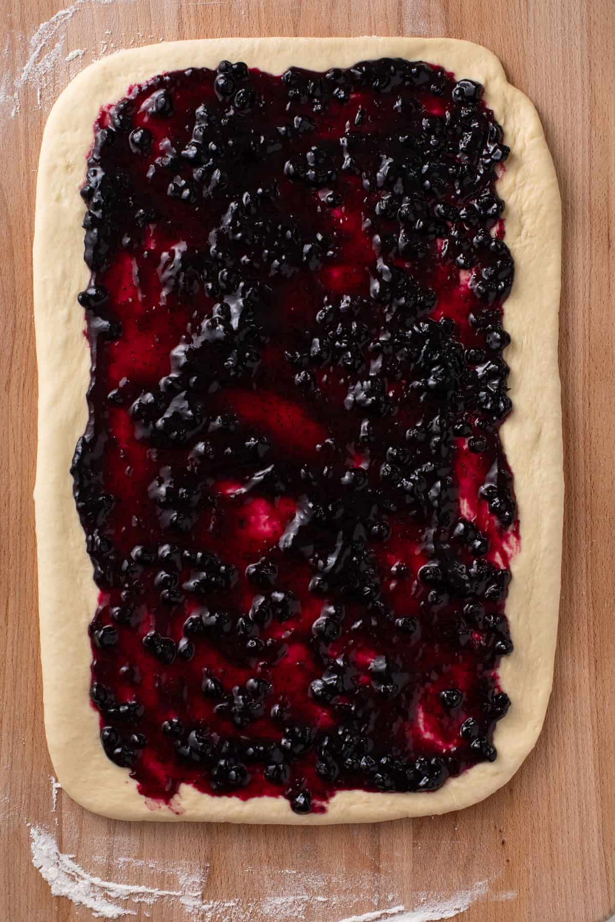 Sweet roll dough rolled into a rectangle on a wooden surface and spread with blueberry preserves.