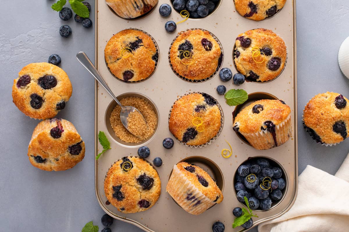 Cooled lemon blueberry muffins arranged in a muffin tin along with fresh blueberries.