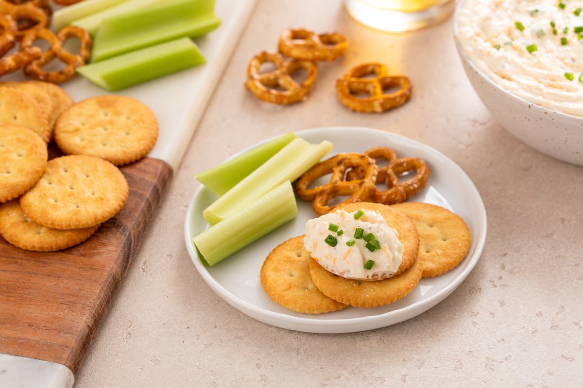 White plate on a countertop, holding crackers with beer cheese dip, pretzel twists, and celery sticks.