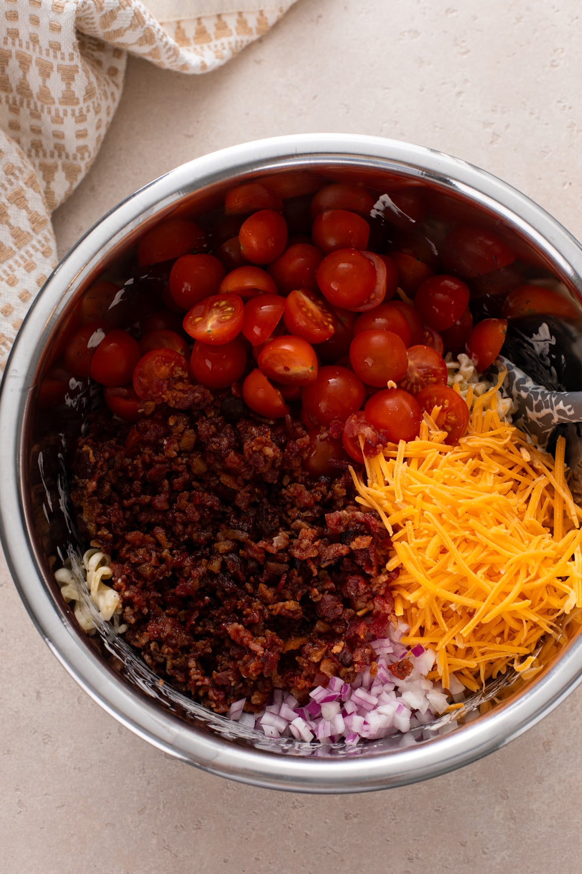 Halved cherry tomatoes, shredded cheese, crumbled bacon, and diced red onion added to a metal bowl.