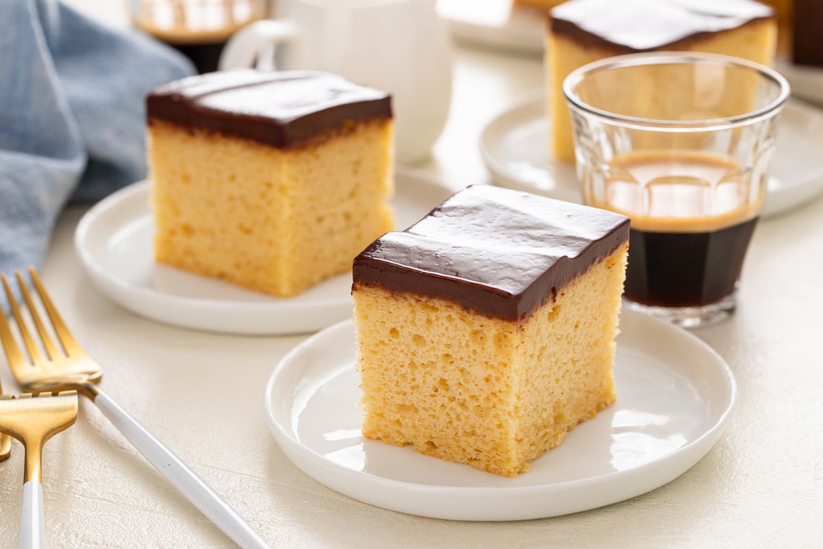 Two slices of yellow sheet cake with chocolate frosting, both on white plates with a glass of espresso in the background.