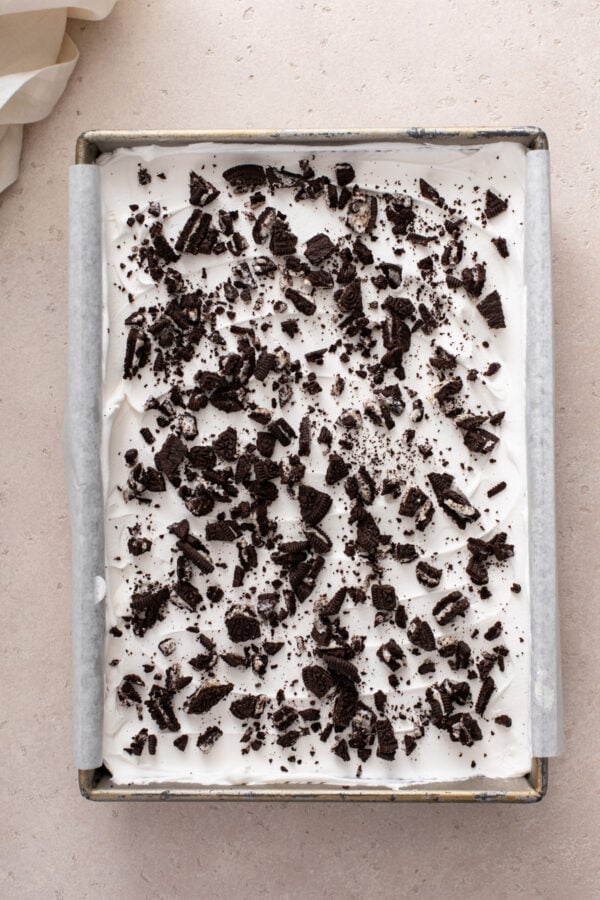 Layered chocolate pudding dessert assembled in a parchment-lined baking pan, topped with cool whip and crushed oreo cookies.