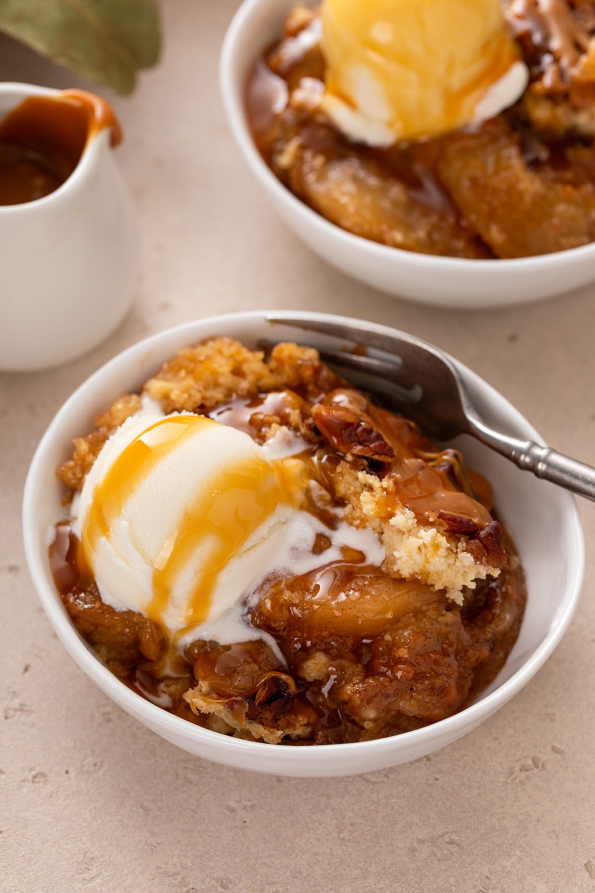 Fork in a bowl of caramel apple dump cake topped with a scoop of vanilla ice cream and caramel sauce.