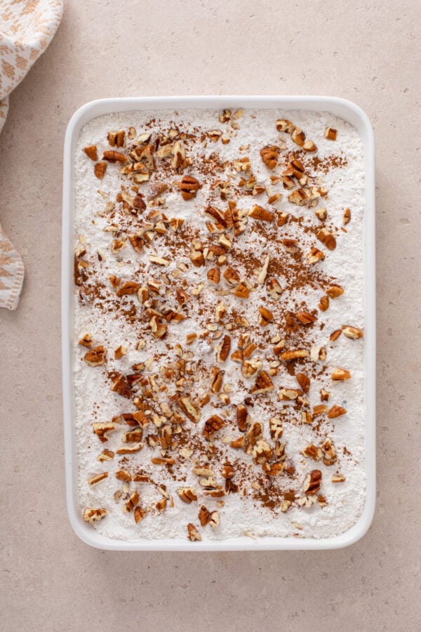 Cake mix and chopped pecans spread on top of the filling for caramel apple dump cake in a white pan.