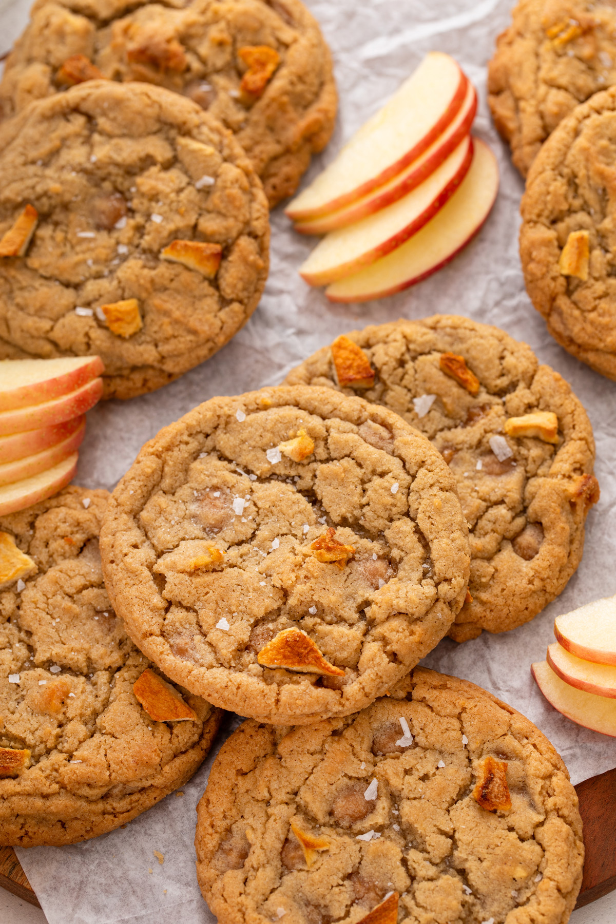 Close up view of caramel apple cookies scattered on parchment paper amongst fresh apple slices.