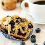 Blueberry lime muffin cut in half on a white surface in front of a cup of coffee and fresh blueberries