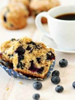 Blueberry lime muffin cut in half on a white surface in front of a cup of coffee and fresh blueberries